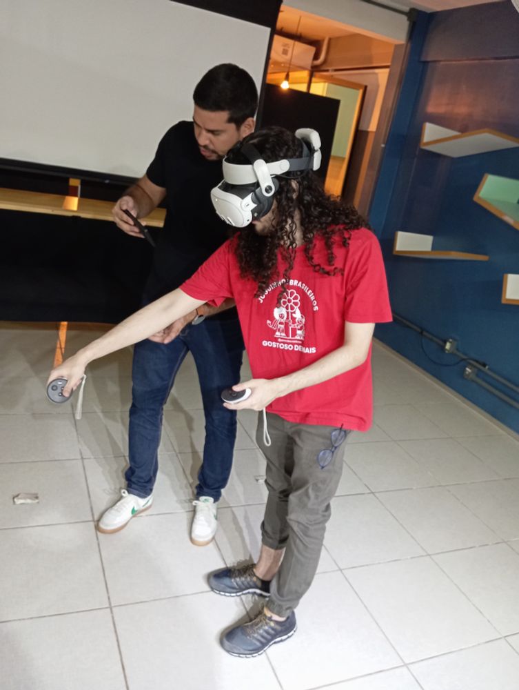 me, with long dark brown hair, red shirt, grey pants and shoes, using VR goggles, stretching my arm trying to grab something with the controllers.