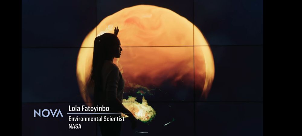 Lola Fatoyinbo in front of a visualization of Global Carbon Dioxide shown in orange playing on a large tiled display.