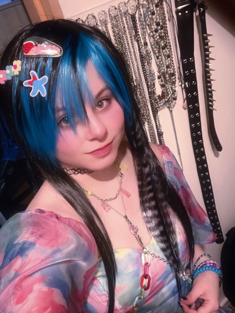 A selfie of me in a colorful dress & jewelry. My hair is black & blue in a scene style with a black & grey raccoon tail. Three cute hair clips are in my hair