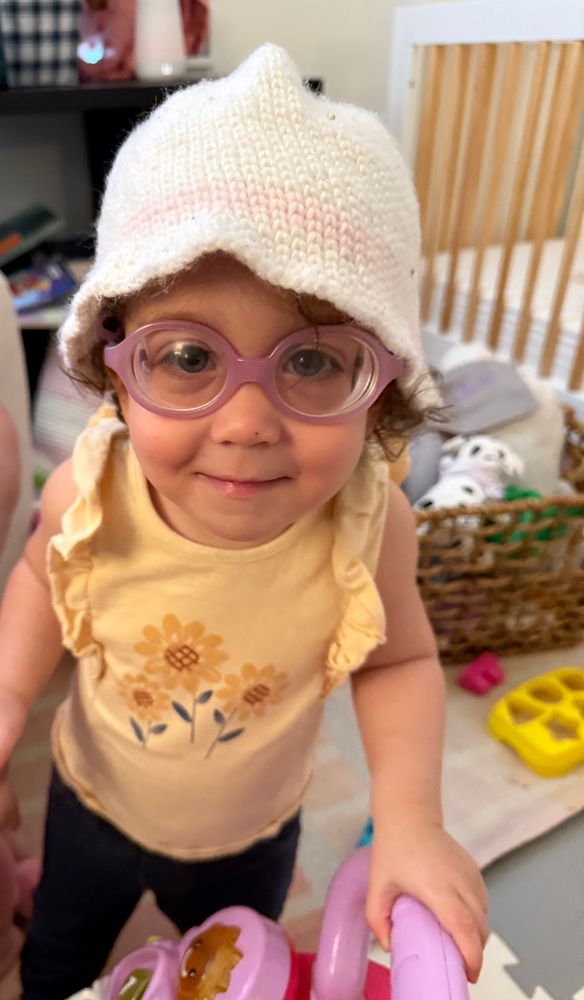 My toddler daughter in a white shapeless knit hat and purple glasses