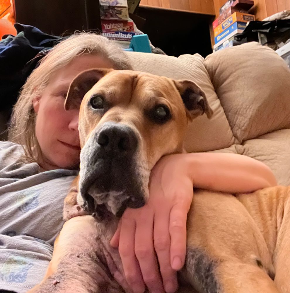 Woman in an overstuffed chair. She is mostly hidden by the large brown boxer dog sprawled on her. She has an arm around the dog’s shoulders and the dog is looking at the camera. 