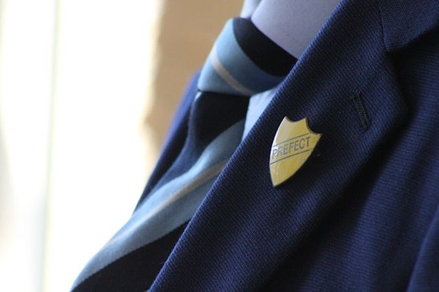 A close-up shot of someone wearing a navy blazer with a yellow, enamel crest-shaped pin badge saying “prefect”. The person is also wearing a white shirt and a white, pale blue and navy blue striped tie. You can only see the clothes, not the person wearing them. 
