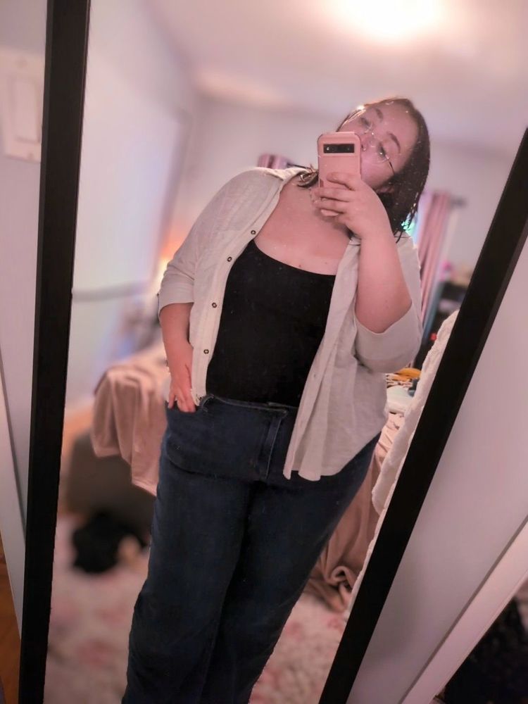 Dani taking a mirror pic while standing in front of her bed. She is wearing dark jeans, a navy blue tank top, and a loose white button up shirt that is left open. Rare sight of her hair down, not in pigtails