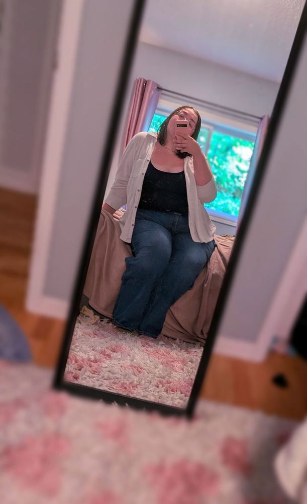 Dani taking a mirror pic while sitting on her bed. She is wearing dark jeans, a navy blue tank top, and a loose white button up shirt that is left open. Rare sight of her hair down, not in pigtails