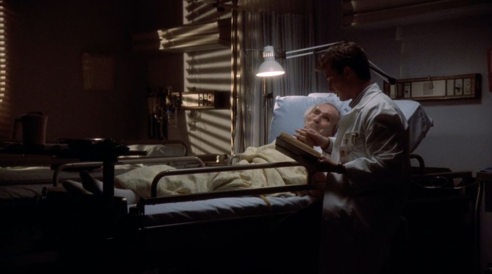 Estamos em quarto de hospital escuro, a única iluminação vem de uma luminária branca posicionada acima da maca de hospital, onde está deitado um senhor idoso. Ao seu lado está sentado John Carter, ele é um homem branco e tem cabelos castanhos. Está usando um jaleco branco e com a mão esquerda segura um livro, que está lendo, e com a mão direita segura a mão do senhor que está na maca.