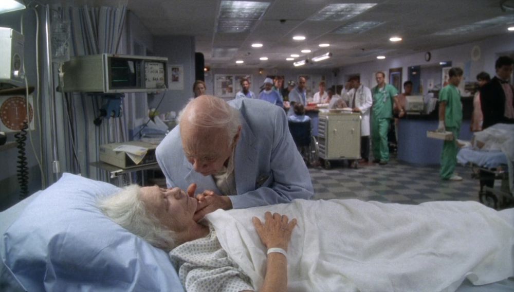 Imagem da série de televisão ER. Vê-se um espaço de hospital, branco e bem iluminado, em plano longo. O chão é de lajotas brancas e azuis. Próximo da cama, está uma maca, com uma senhora idosa branca e de cabelos brancos deitada tapa com um lençol também branco. Ao seu lado, está o seu marido, também um idoso branco, que está segurando sua mão enquanto se curva sobre a maca. Ao fundo, estão vários médicos e enfermeiras observando a cena.