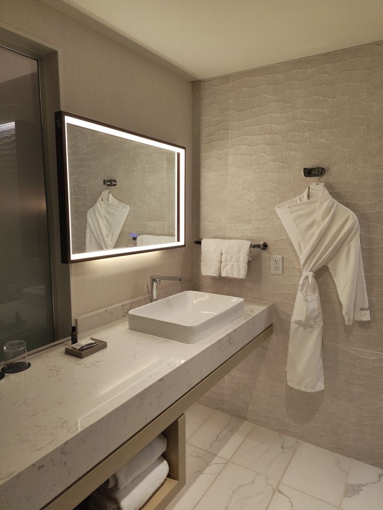 A modern and clean hotel bathroom vanity. The floor and countertop appears to be white marble. A nicely folded bath robe hangs on the wall.