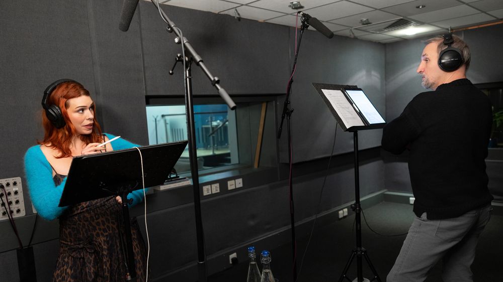 Billie Piper and Christopher Eccleston recording the Ninth Doctor Adventures audio dramas in a studio. They are both wearing headphones and standing in front of mics and lecterns. Billie, left, is looking at Christopher and holding up an iPad stylus. Christopher is looking back attentively. 