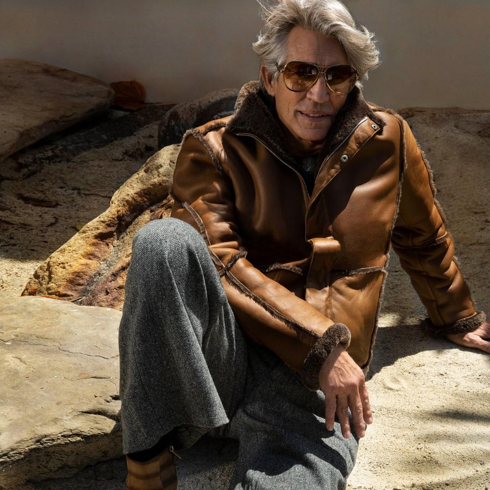 Eric Roberts, looking effortlessly cool in sunglasses. He is sitting on a rock in grey woollen slacks and a tan leather jacket with fur trim. 