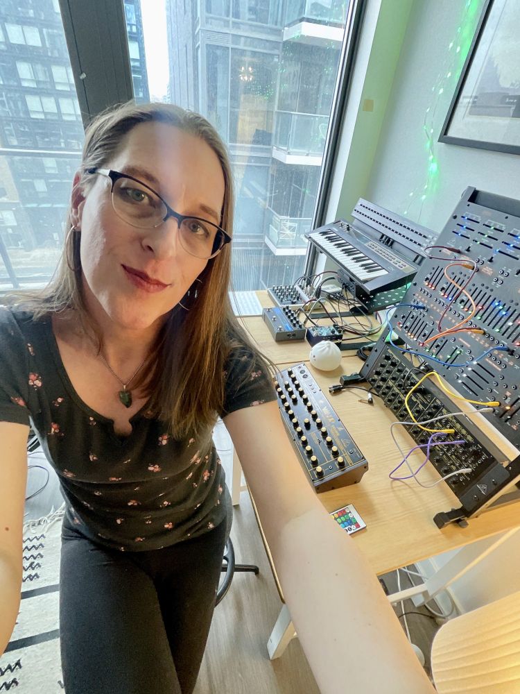 trans woman in front of a table of synthesizers and music gear. She's happy