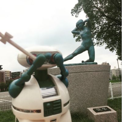 Toy Holiday photo of God Jesus Robot looking embarrassed in front of a statue, even though it is clothed.
