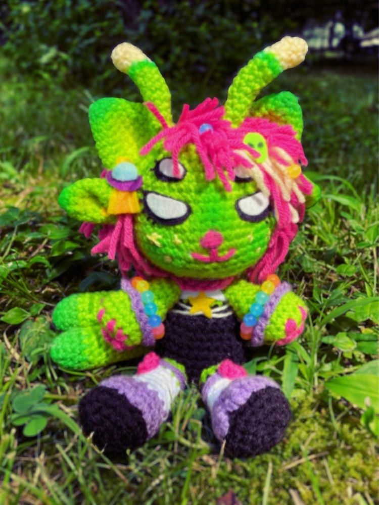 a crocheted green and pink alien plush rests in grass