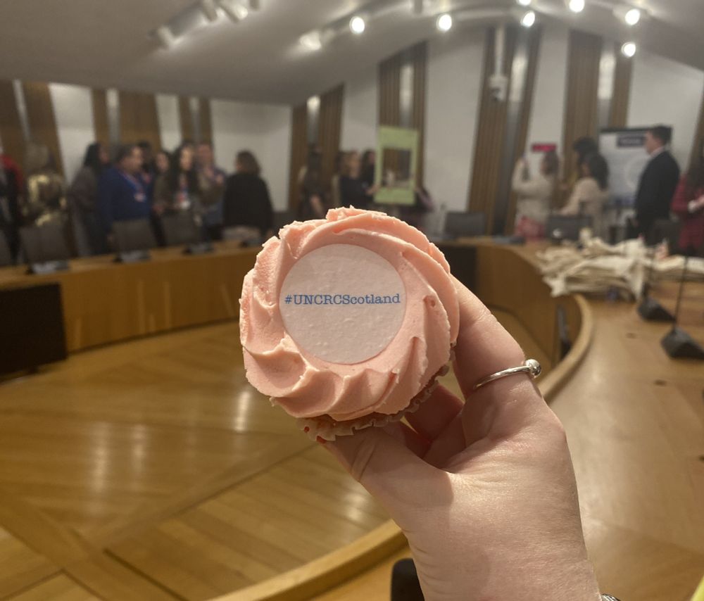 Photo of cupcake and Scottish Parliament room in the background with people. #UNCRCScotland on top. Photo.