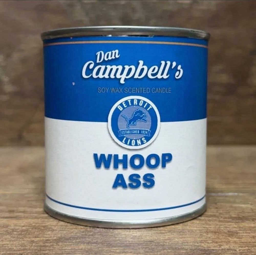This image shows a product labeled as "Dan Campbell's WHOOP ASS," designed to look like a can but is actually a soy wax scented candle. It features the Detroit Lions logo, making it a fun nod to the team's head coach, Dan Campbell, and their fighting spirit.

