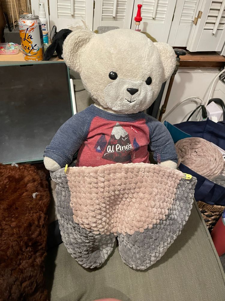 a teddy bear (alex) standing in the legs of an in process amigurumi totoro.
