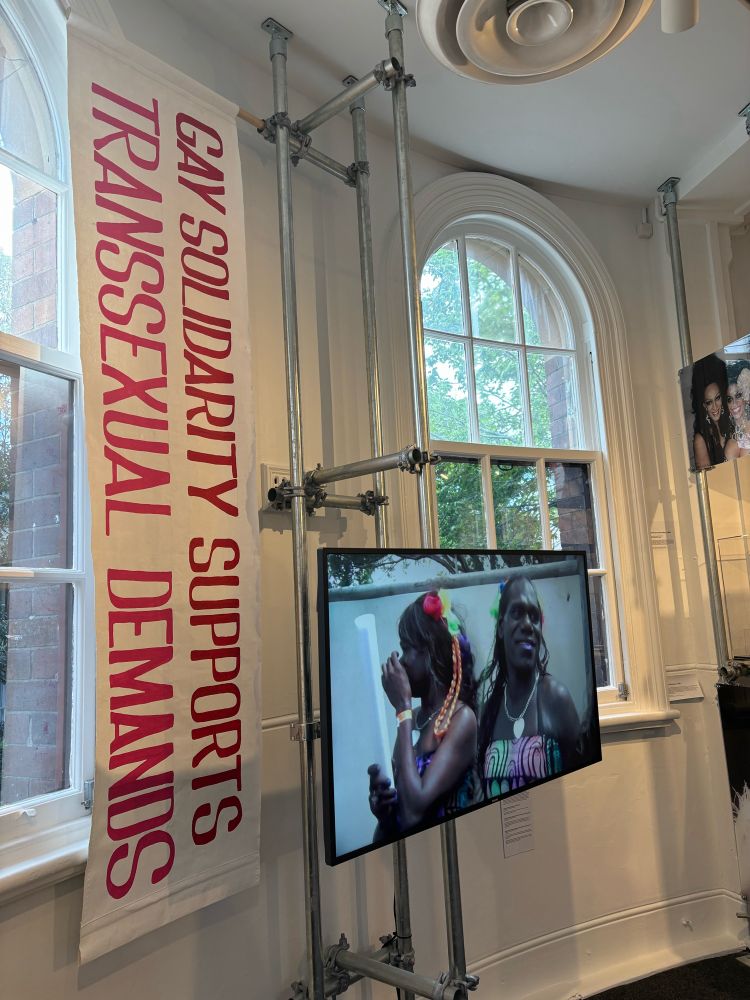 “Gay Solidarity Supports Transsexual Demands” is written on a banner next to a tv screen showing clips of First Nation trans people at Sydney Qtopia museum