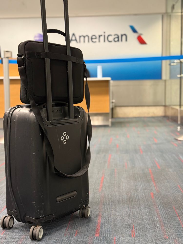 Photo of a Bluesmart carry-on luggage bag, handle extended. A laptop bag rests on top of it, looped around the handle. In the background, the American Airlines logo is visible. Location is inside of an airport. 