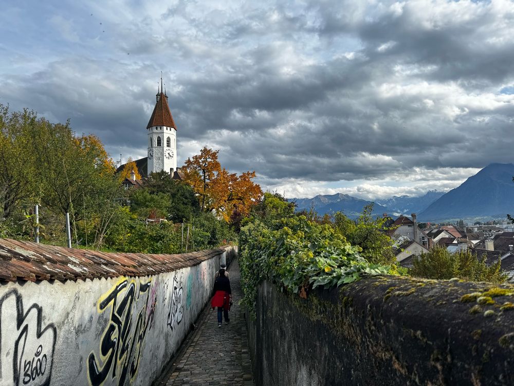 Small street of old town Thun under a partially cloudy blue sky. We see paint tags on a wall and the old town church in the distance