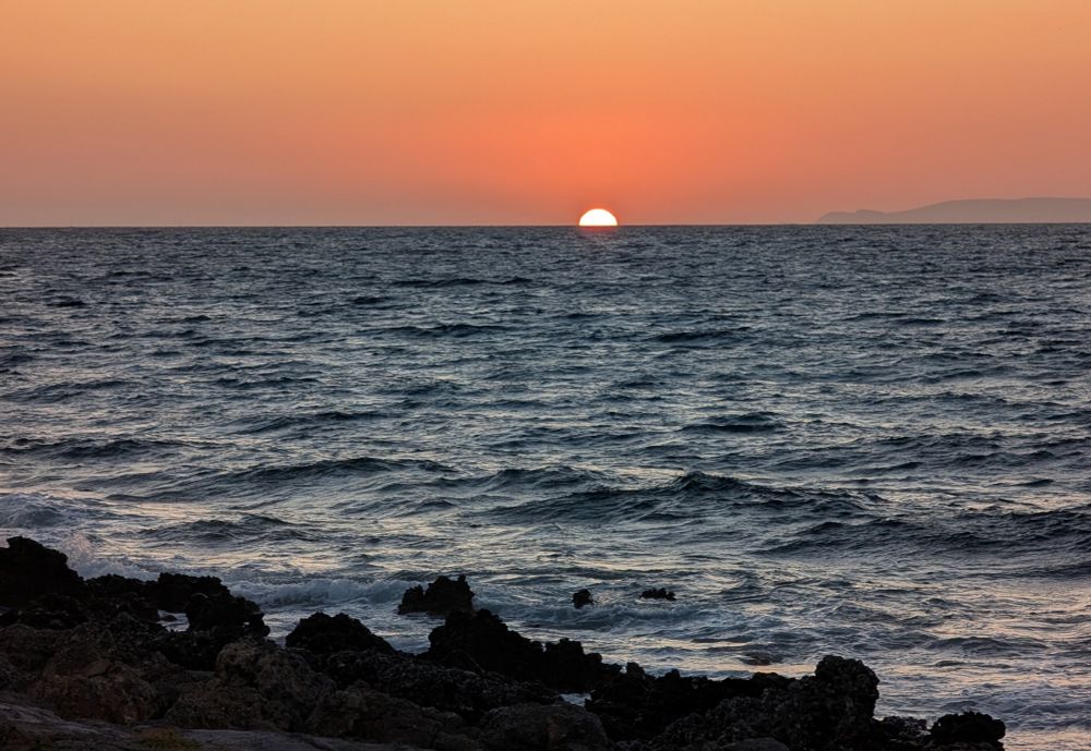 le demi disque jaune du soleil à l'horizon sur une mer calme. le ciel est un dégradé d'oranges et au premier plan se découpent des rochers sombres. 
