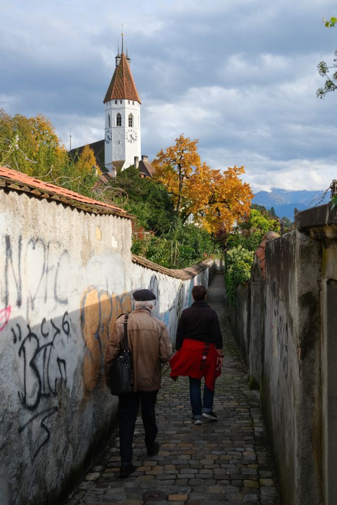 Two persons walking down a narrow street. We see the tip of the church over them