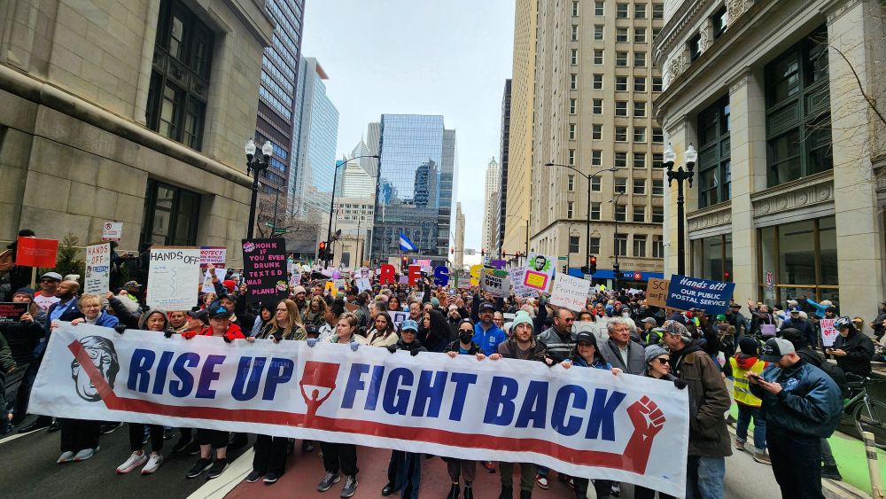 A photo of a large protest in Chicago. Hundreds of people in the street are holding signs. A large banner at the front reads "rise up fight back"