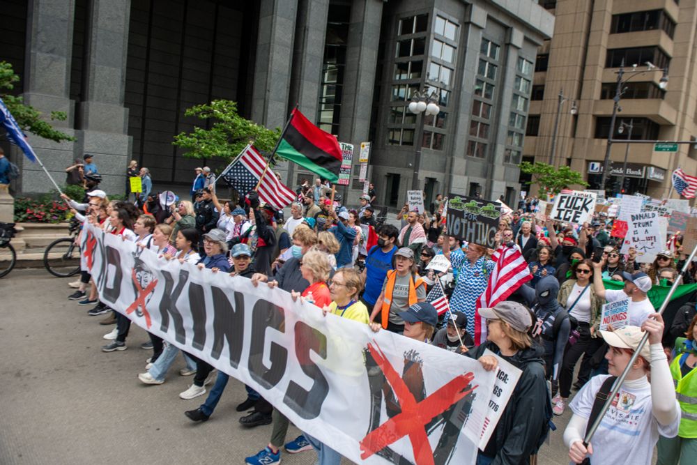 A photo of a large street demonstration in Chicago. At the front many people are holding a large banner that reads "NO KINGS" with several crowns that have red X's through them. Many people are marhcing behind the banner, holding signs and flags. 