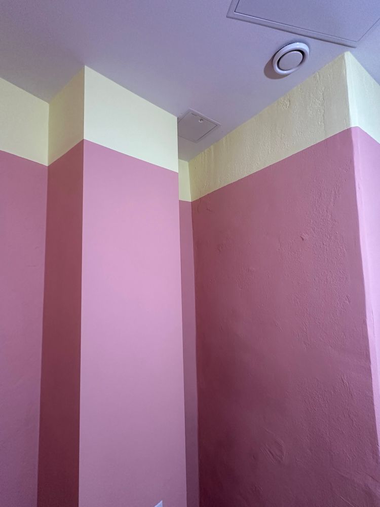 Corner of the backroom with pink walls that have a highlighter yellow slither of color at the top of the roof line.