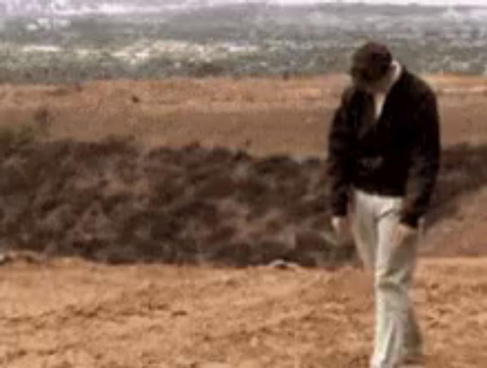 a man in a black jacket and white pants is standing on a dirt field .