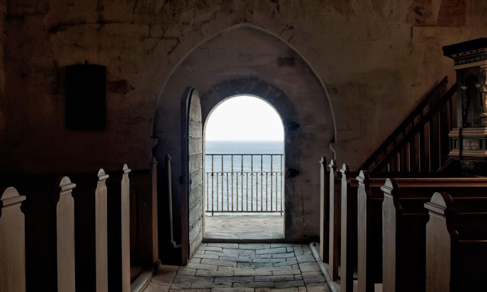 Looking down the worn, stone aisle of a small church with pews either side. There is a view of the ocean through the arched door at the end of the aisle