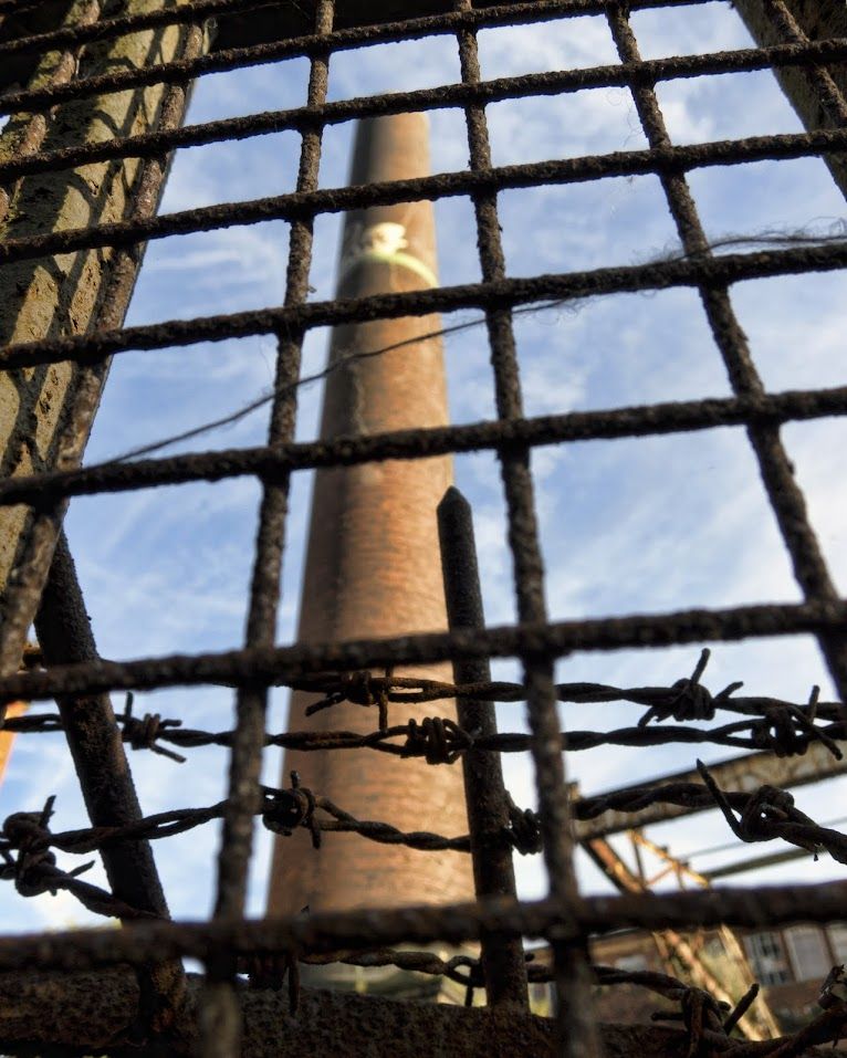 Looking through rusty railings and barbed wire up to a factory chimney 