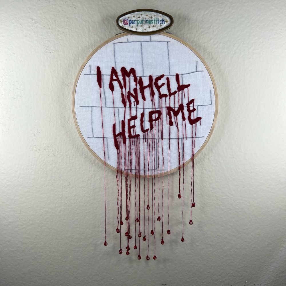Picture of an embroidery depicting the “I am in hell, help me” words written in blood on a wall from Hellraiser II. The dripping blood is represented by strands of floss hanging off of the hoop weighed down with small glass red beads. The stitcher’s handle (@purpurinastitch) is visible on a needleminder on the hoop.