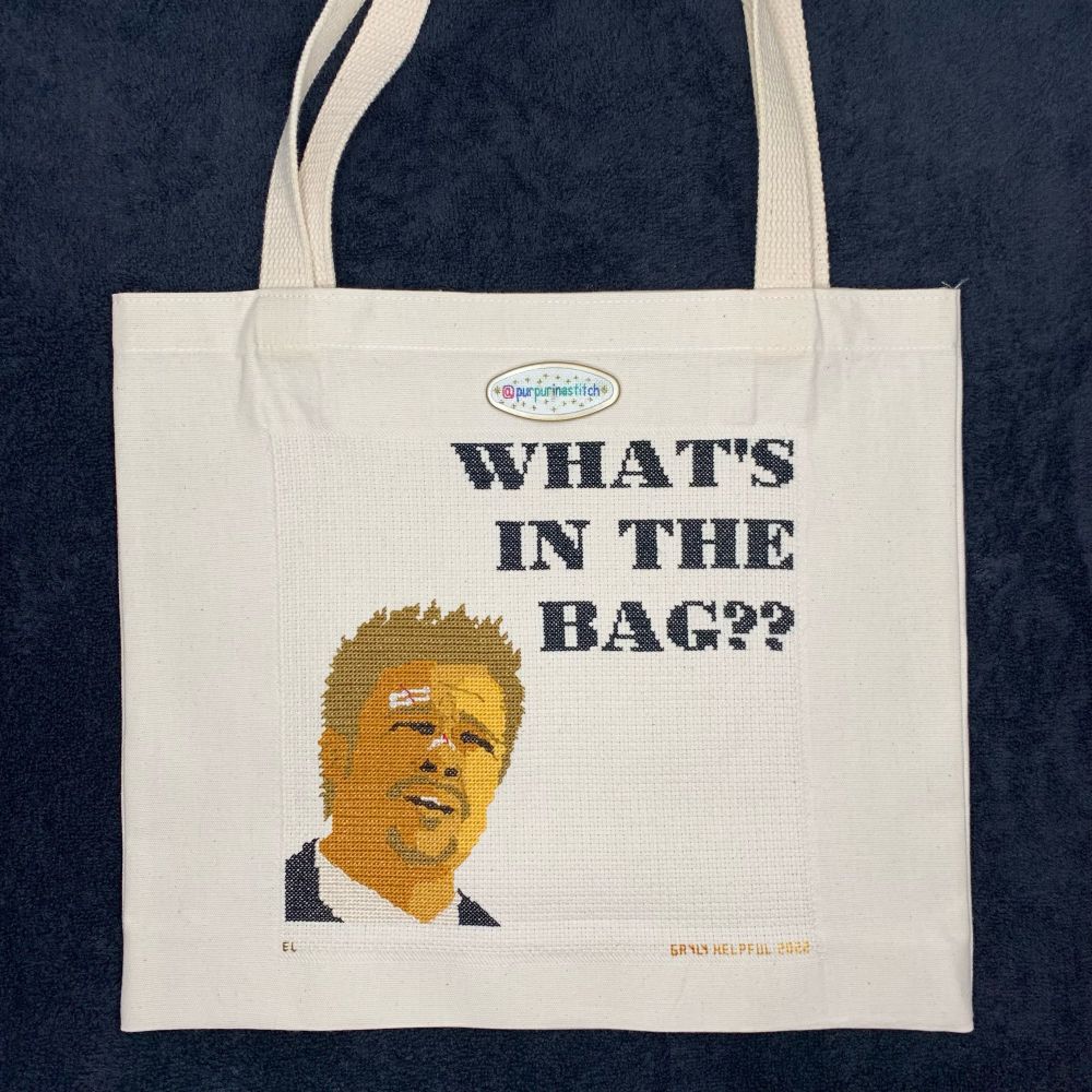 Picture of a cross stitched canvas tote bag depicting Brad Pitt’s character from the end of the movie Seven. The text “What’s In The Bag??” is stitched in large block letters above the character’s head. The stitcher’s handle (@purpurinastitch) is visible on a needleminder on the fabric.