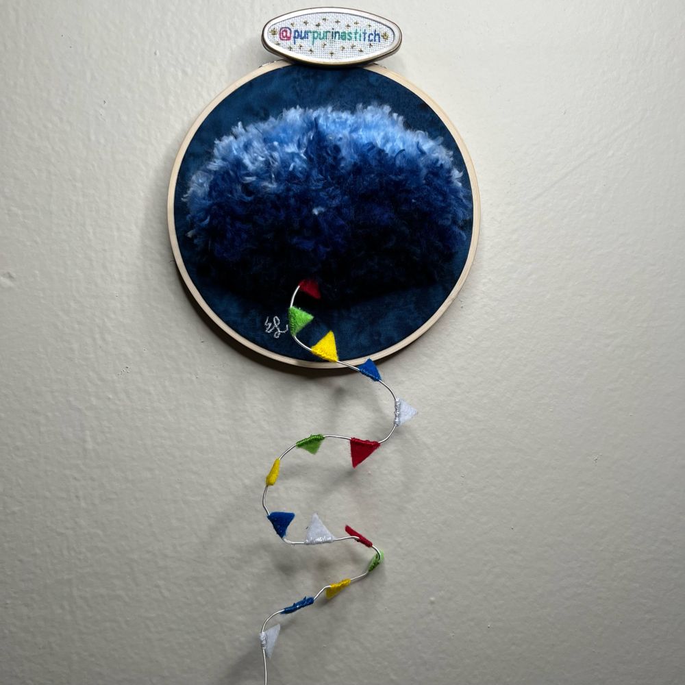 Picture of an embroidered cloud similar to the one depicted on the NOPE movie posters. A tail of flags made out of felt is hanging out of the cloud and off of the embroidery hoop. The stitcher’s handle (@purpurinastitch) is visible on a needleminder on the hoop.