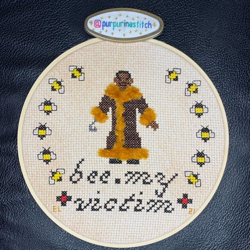 Picture of a completed cross stitch featuring Candyman over the quote/pun “bee my victim,” referencing a line in the first Candyman movie. There is a wreath of bumblebees. Candyman’s coat has turkey work stitching on the trim to emulate fur. The stitcher’s handle (@purpurinastitch) is visible on a needleminder on the fabric