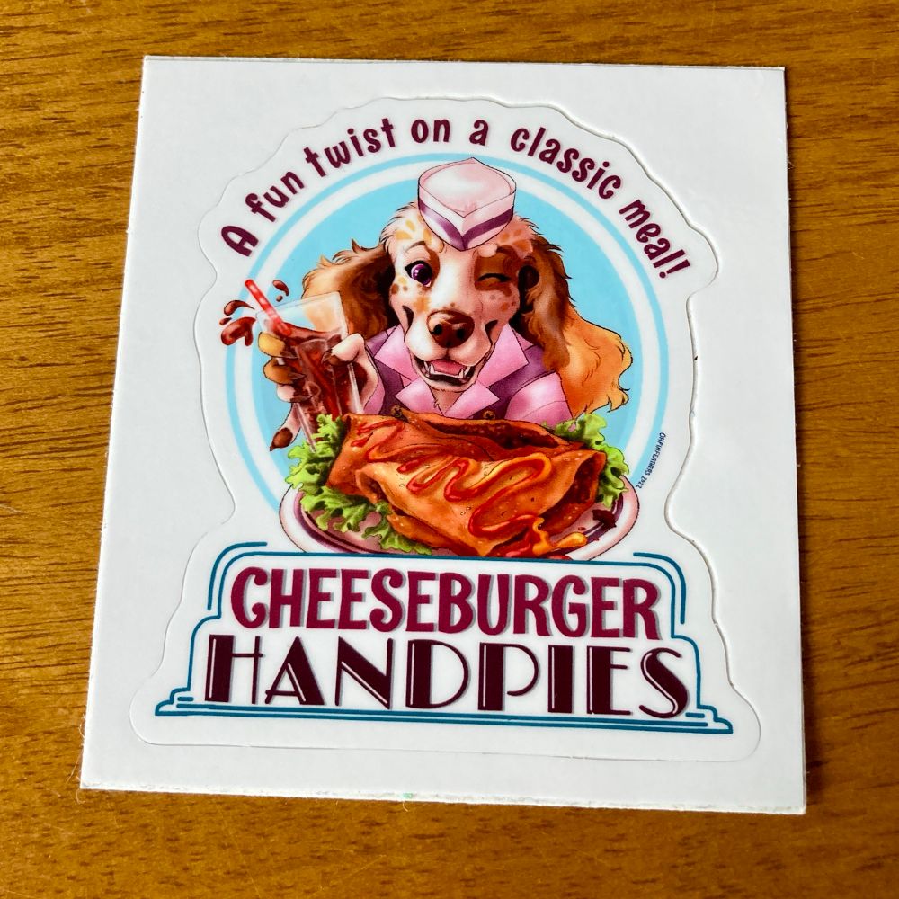 Sticker of Buttercup the baking spaniel. She is holding a plate with a pastry covered in condiments, on a bed of lettuce, and a glass of soda in the other hand. The text around the sticker reads: “A fun twist on a classic meal! Cheeseburger Handpies”