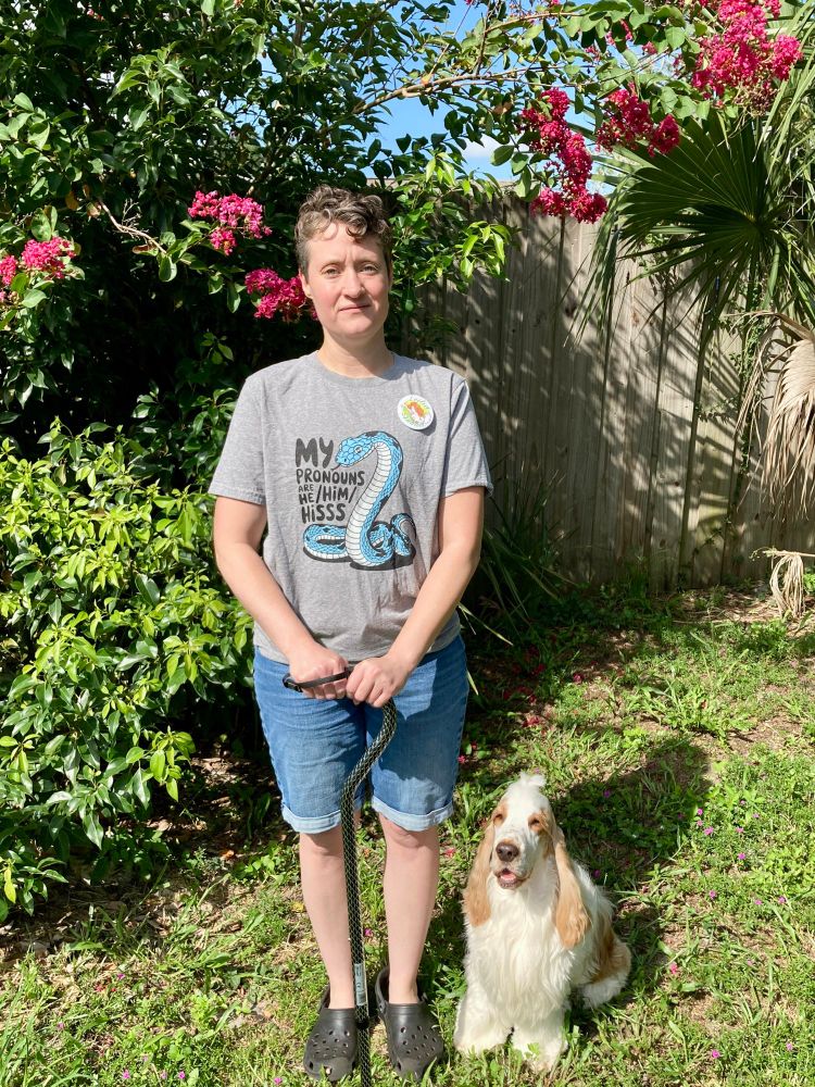 A photo of me standing in my backyard. I am wearing a grey shirt with a blue snake on it. The text reads “My pronouns are he/his/hisss”. There is a button on my shirt featuring art of my character Leilia Spaniel. I am holding a black and silver cane in front of me.

My English cocker spaniel Basil is sitting next to me(he will not be joining me at Chomp, lol).