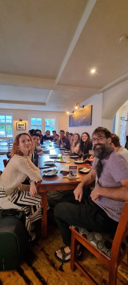 About 15 people sit together at a long wooden table in a restaurant, smiling toward the camera