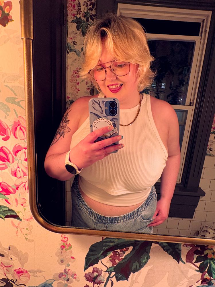 A blonde nonbinary person takes a mirror selfie. They wear a white cropped tank with high waisted light wash jeans and a short gold chain necklace.