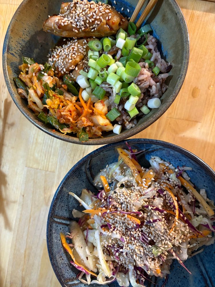 Two bowls on a wooden surface. One holds chicken, wild rice, kimchi with sesame and green onion. Another has a multicolour cabbage sesame slaw. The bowls are blue pottery.