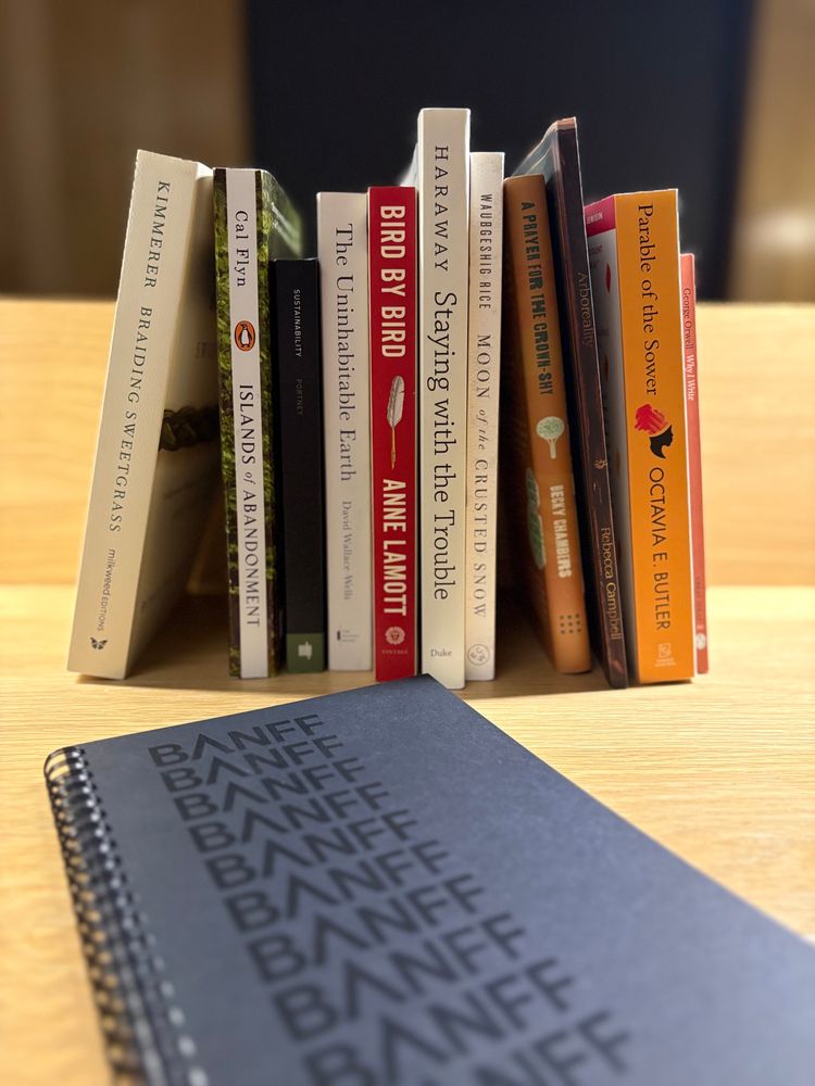 A photo of a line of books (selections from Donna Haraway, Octavia Butler, Becky Chambers, and climate books both fiction and nonfiction) on a light wooden desk, with a notebook before them with all caps BANFF cascading down its front.