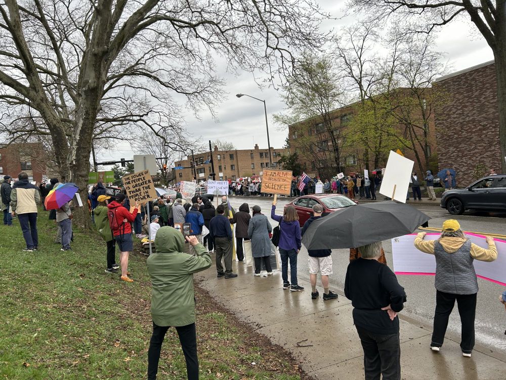 People standing along Fifth Ave in Pittsburgh on a wet, rainy evening with protest signs and umbrellas as cars drive by. 