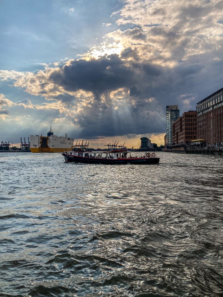 Blick auf den Hamburger Hafen und das Wasser der Elbe. Eine Wolke hat sich gerade vor die Sonne geschoben und sorgt für spannende Lichtstimmung. Auf dem Wasser ist eine Barkasse zu sehen, im Hintergrund fährt ein größeres Frachtschiff den Fluss hinunter.