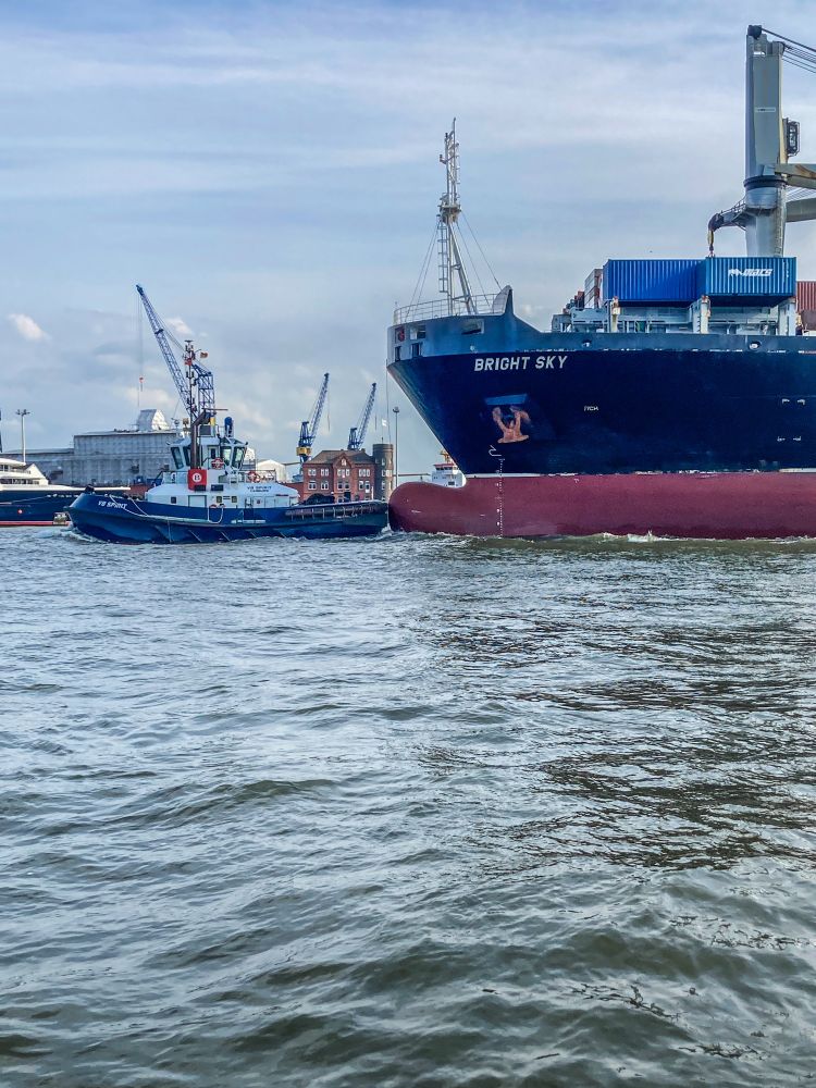 Das Frachtschiff Bright Sky fährt in den Hamburger Hafen ein. Zu sehen ist nur der Bug des Schiffes, links daneben ein Schlepper. Der Schlepper drückt bremsend gegen den Wulstbug des Schiffes, der mangels Tiefgang deutlich über der Wasseroberfläche herausragt. 
Im Vordergrund das Wasser der Elbe, darüber blauer Himmel.