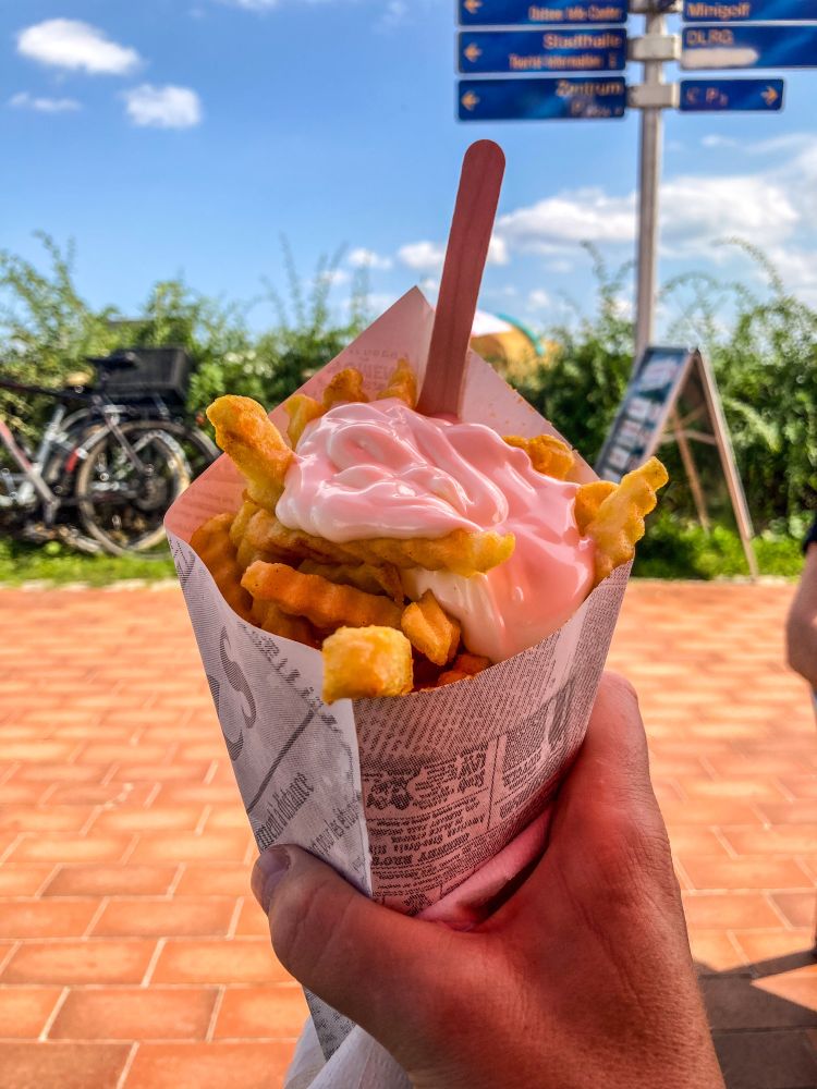 Meine Hand hält eine Portion Pommes mit Mayonsise. Im Hintergrund die Strandpromenade von Eckernförde mit einem rot gepflasterten Gehweg. Sonnenlicht, blauer Himmel.