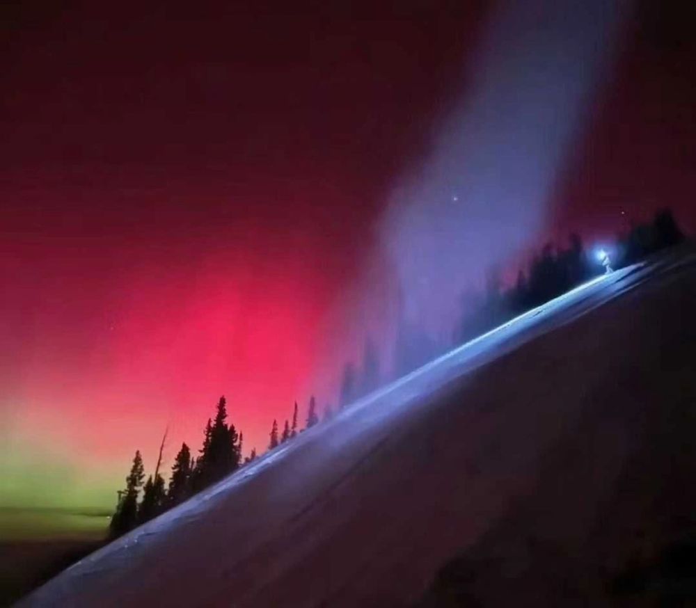 Aurora borealis, reds and greens, in the sky. In the foreground, an icy slope that will be a World Cup downhill course in a few weeks. The light on the right is a snowmaking gun.