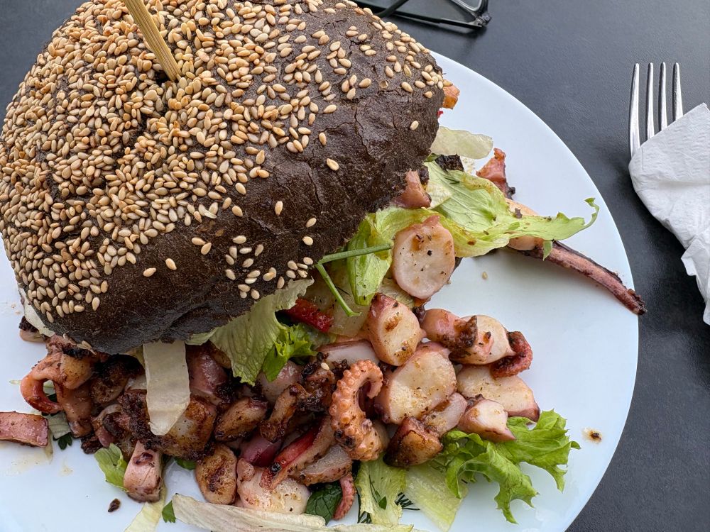 Der Black Pearl Burger. Octopusburger mit dunklem Bun und viel Octopus auf weißem Teller. 