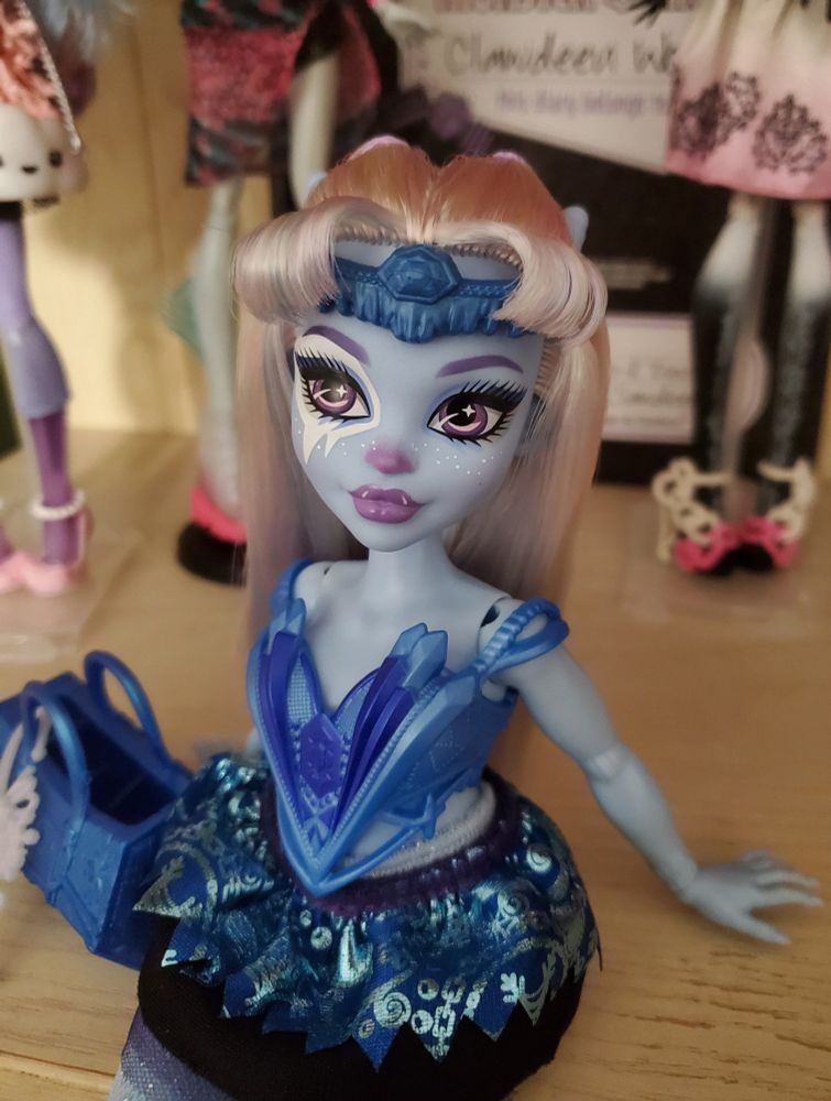 a 3/4 shot image of Abbey Bominable monster mysteries dressed in her Ballerina skirt, corset snap on top, and a new hair style. white, lavender, and ice blue saran hair is styled into a partially up partially down look. her long straight hair is pulled into two mini pigtails between her horns, and she has curled bangs split down the center to showcase her blue circlet, which the forehead skimming part of her headset cut off.