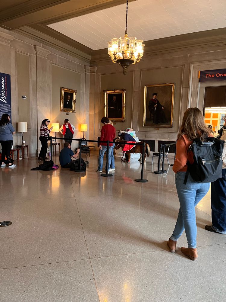 A mini horse wearing festive gear is greeted by a line of patrons in the library gallery. 
