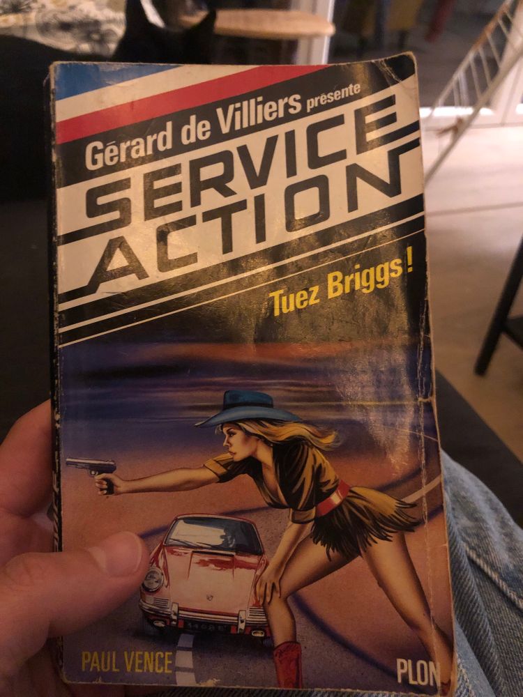 Couverture du livre « Gérard de Villiers présente : Service Action, Tuez Briggs ! ». Un gros titre dégueulasse en bandeau, et dessous, un montage ignoble d’une Datsun rouge avec (?!) devant, une cow-girl texane penchée en train de tirer au pistolet. Un vrai régal visuel. 