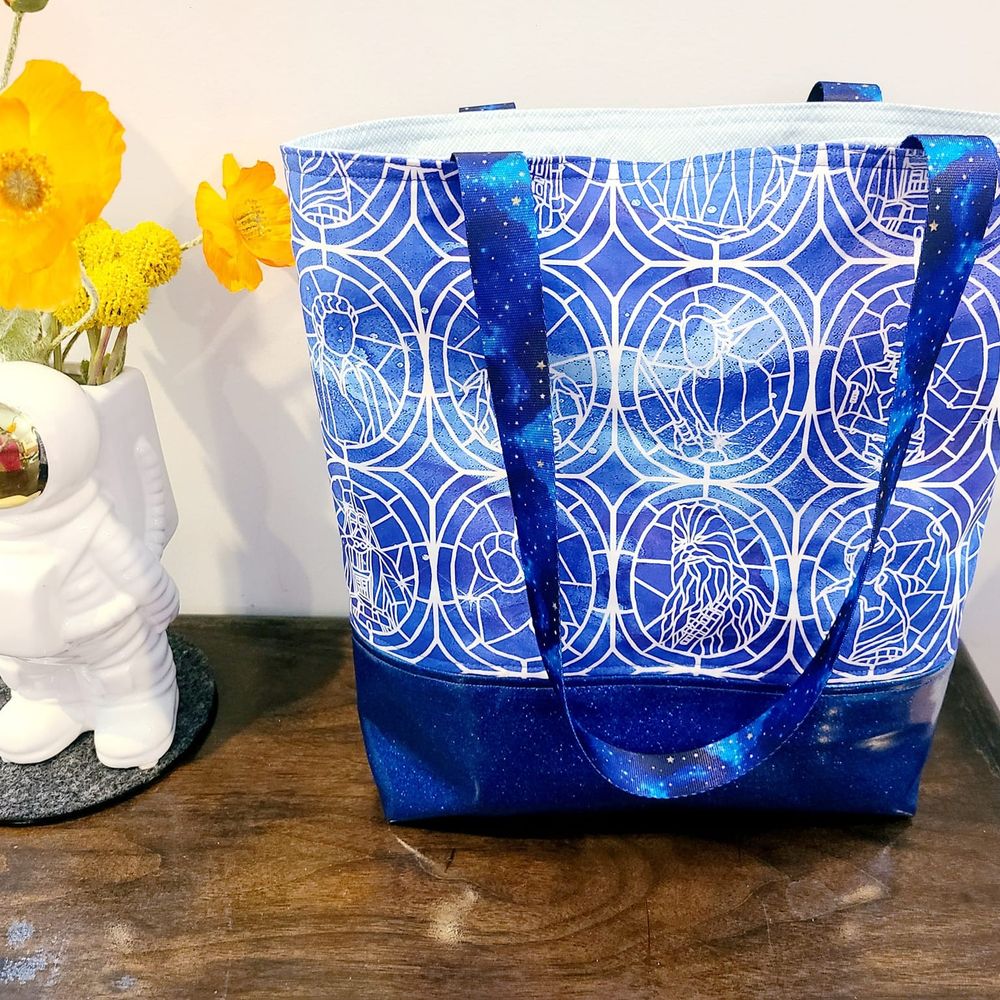 On the left is a vase shaped like a 1960s astronaut with fake orange poppies in it. On the right is a blue tote bag. It has a glittery bottom. The main fabric is stained glass style and shows characters from the original Star Wars. The straps have a blue space print on them. Everything is sitting on a brown wooden table.